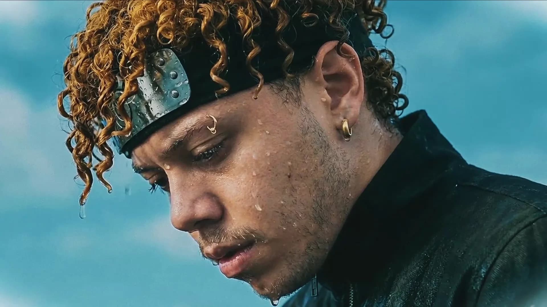 Close on a man's face in cold daylight, head bowed, water running off his curls, pale sky behind him.